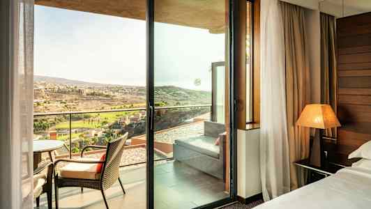 Hotellihuone, josta on näkymä lasioven kautta parvekkeelle, jossa on tuoleja ja kaukainen maisema ulkona. (Tekoälyn luoma teksti)