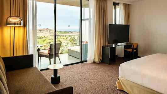 Hotellihuone, jossa istuinryhmä, sänky, televisio ja näkymä parvekkeelle, jolta avautuu maisema. (Tekoälyn luoma teksti)