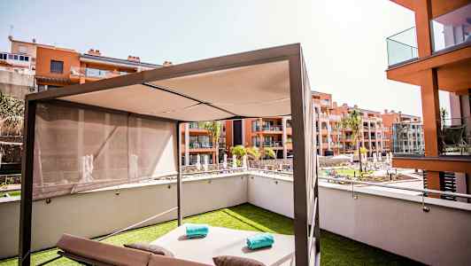 Moderni aurinkovuode aurinkovarjon kanssa nurmikolla, jota ympäröivät asuinrakennukset ja uima-allas. (Tekoälyn luoma teksti)