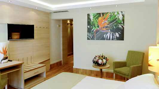 Hotellihuone, jossa on sänky, vihreä tuoli, kukkakimppu pöydällä ja seinäkuva oranssista kukasta. (Tekoälyn luoma teksti)