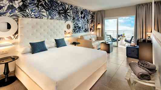 Hotellihuone, jossa suuri sänky, istuinryhmä ja parveke näkymällä. (Tekoälyn luoma teksti)