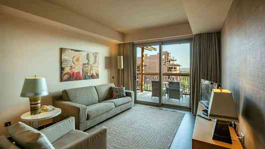 Olohuone, jossa on sohva, nojatuoli ja sohvapöytä, televisio huonekalulla sekä lasiovi, joka johtaa parvekkeelle. (Tekoälyn luoma teksti)