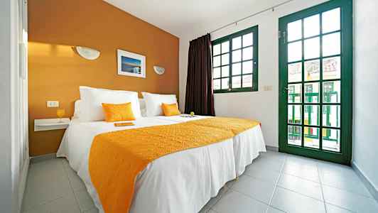 Makuuhuone, jossa on parivuode, valkoiset ja oranssit tekstiilit, sekä suuret vihreäreunaiset ikkunat. (Tekoälyn luoma teksti)