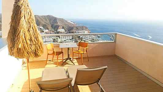 Näkymä terassilta, jossa on aurinkovarjo, kaksi tuolia ja pöytä sekä aurinkotuolit merinäköalalla. (Tekoälyn luoma teksti)