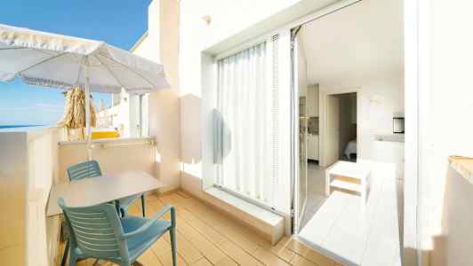 Parveke, jossa on pöytä ja tuolit, aurinkovarjo ja näkymä merelle. Ovi johtaa valoisaan sisätilaan, jossa on huonekaluja. (Tekoälyn luoma teksti)