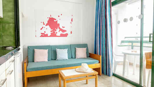 Olohuone, jossa on turkoosi sohva, värikkäitä tyynyjä, pöytä ja hattu. Ikkuna, josta näkyy parveke. (Tekoälyn luoma teksti)