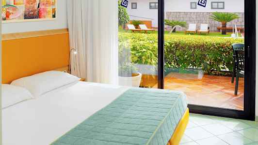 Hotellihuone, jossa sänky suuren lasi-ikkunan edessä, ikkuna avautuu vehreälle puutarhalle. (Tekoälyn luoma teksti)