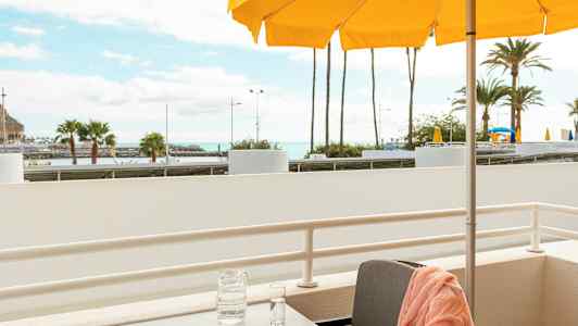 Parveke, jossa pöytä, oranssi pyyhe tuolilla, karahvi ja lasi pöydällä sekä taustalla keltaisia aurinkovarjoja ja palmuja. (Tekoälyn luoma teksti)