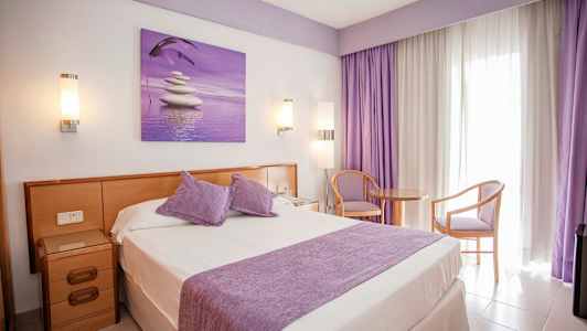 Hotellihuone, jossa parivuode, violetteja yksityiskohtia ja istumapaikka ikkunan vieressä. (Tekoälyn luoma teksti)