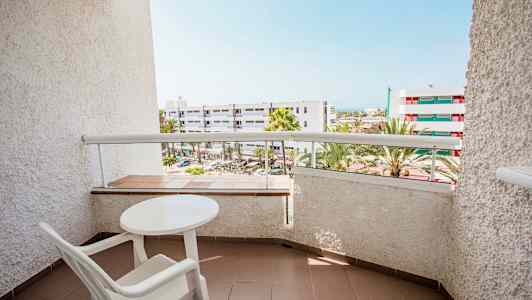 Parveke kuva, jossa pöytä ja tuoli, näkymä rakennuksiin ja palmuihin. (Tekoälyn luoma teksti)