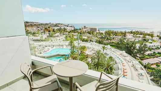 Parveke jossa kaksi tuolia ja pyöreä pöytä sekä näkymä hotellialueelle uima-altaalla ja merelle. (Tekoälyn luoma teksti)