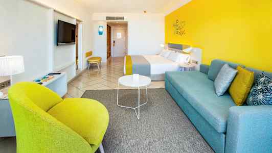 Modernisti sisustettu hotellihuone, jossa värikkäitä yksityiskohtia, kuten keltainen tuoli ja sininen sohva. (Tekoälyn luoma teksti)