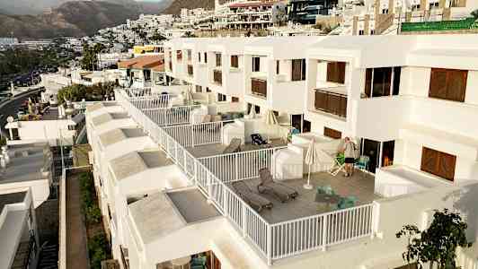 Kuva rivistöstä moderneja valkoisia huoneistoja, joissa on suuret yksityiset parvekkeet, sisustettu tuoleilla ja aurinkotuoleilla, mäkisessä ympäristössä vuorten taustalla. (Tekoälyn luoma teksti)