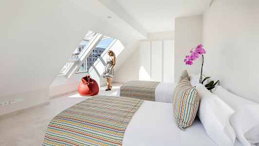 Valoisa huone, jossa on kaksi sänkyä, värikkäitä tyynyjä, punainen istuinpussi ja suuri viisto ikkuna. (Tekoälyn luoma teksti)