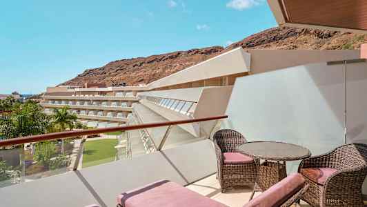 Terassi, jossa aurinkotuoleja ja huonekaluja, näkymä hotellirakennuksiin, palmupuihin ja vuoriin. (Tekoälyn luoma teksti)