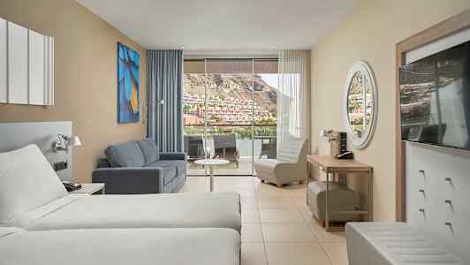 Hotellihuone kahdella sängyllä, sohvalla, nojatuolilla ja suurella parvekkeella, josta näkymä vuoristoiselle alueelle. (Tekoälyn luoma teksti)