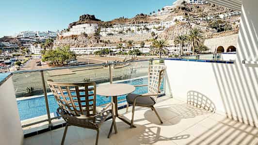 Parveke, jossa kaksi tuolia ja pöytä, näkymä altaalle, rakennuksille ja vuorille. (Tekoälyn luoma teksti)