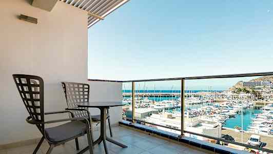 Kuva näyttää parvekkeen, jossa on kaksi tuolia ja pöytä, näkymä satamaan, jossa veneitä. (Tekoälyn luoma teksti)