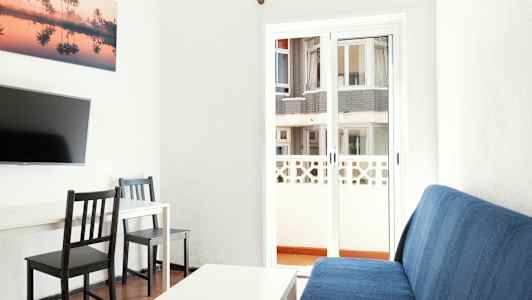 Sisäkuva olohuoneesta, jossa on sininen sohva, valkoinen pöytä, kaksi mustaa tuolia ja televisio seinällä. Lasioven takana näkyy rakennus. (Tekoälyn luoma teksti)