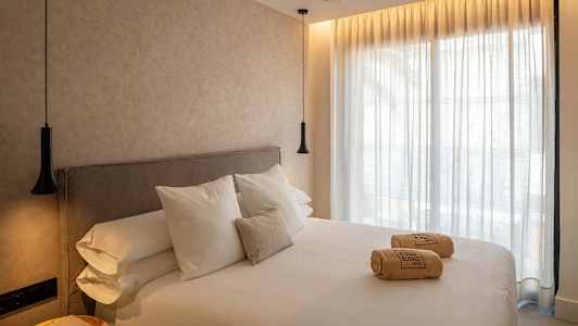 Hotellihuone, jossa on suuri sänky, valkoiset lakanat ja tyynyt. Sängyllä on rullattuja pyyhkeitä. Seinät ja verhot ovat vaaleita ja huoneessa on riippuvalaisimet. (Tekoälyn luoma teksti)