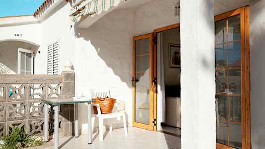 Terassi, jossa on valkoinen rakennus, pöytä ja tuoli sekä taloon johtava ovi. (Tekoälyn luoma teksti)