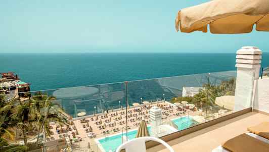 Näkymä terassilta, jossa on pöytä ja tuoleja, kohti uima-allasta, jota ympäröivät aurinkotuolit ja aurinkovarjot, taustalla näkyy meri. (Tekoälyn luoma teksti)