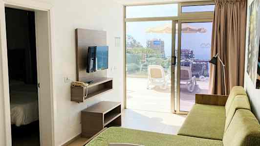 Hotellihuoneen näkymä vihreällä sohvalla ja seinällä olevalla televisiolla, ovi makuuhuoneeseen ja terassi, josta näkyy ympäristö ulkona. (Tekoälyn luoma teksti)