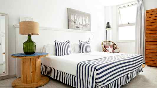 Makuhuone, jossa on raidallisilla tekstiileillä koristeltu sänky, vihreä lasilamppu pöydällä ja rottinkituoli vierellä. (Tekoälyn luoma teksti)