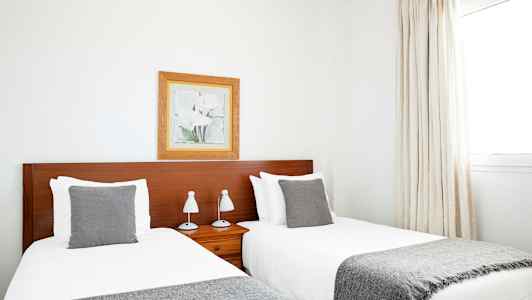 Kaksi yhden hengen vuodetta, joissa harmaat peitot ja tyynyt, sijaitsevat vierekkäin puunvärisen sängynpäädyn kanssa. Kehystetty muotokuva roikkuu seinällä sängynpäädyn yläpuolella. (Tekoälyn luoma teksti)