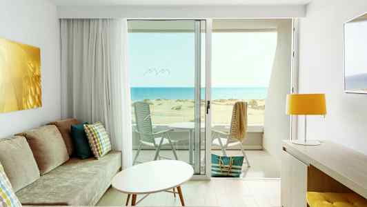 Näkymä valoisasta hotellihuoneesta, jossa on moderni sisustus, aurinkoiselle rannalle ja merelle. (Tekoälyn luoma teksti)