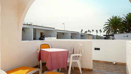 Se näyttää terassin, jossa on aurinkotuoleja ja pöytä tuoleineen. Taustalla on pieniä taloja ja palmuja. (Tekoälyn luoma teksti)