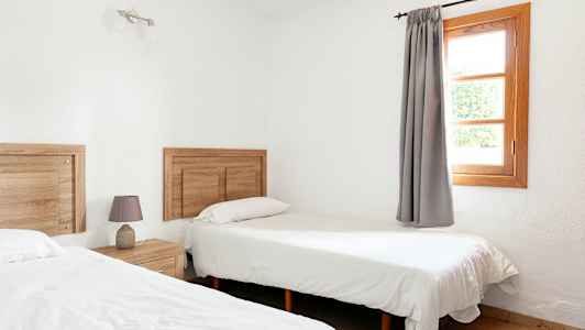 Huone, jossa on kaksi yhden hengen vuodetta, puuaiheita ja ikkuna harmailla verhoilla. (Tekoälyn luoma teksti)