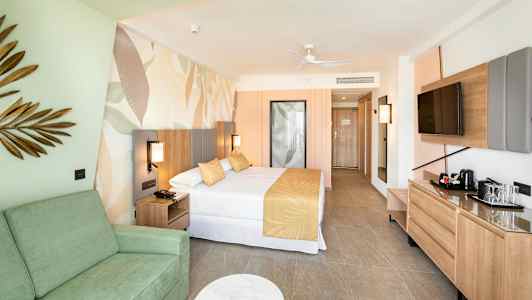 Hotellihuone, jossa sänky, sohva ja televisio. Sisustus vaaleissa väreissä. (Tekoälyn luoma teksti)