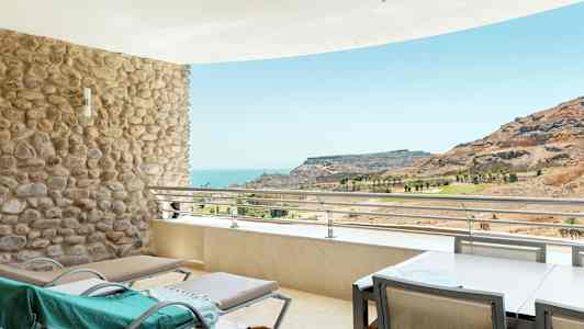 Terassi, jossa aurinkotuoleja, näkymä merelle ja vuoristomaisema. (Tekoälyn luoma teksti)