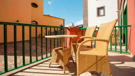 Kaksi punottua tuolia ja pieni pöytä sijaitsevat aurinkoisella terassilla, taustalla värikkäitä rakennuksia. (Tekoälyn luoma teksti)