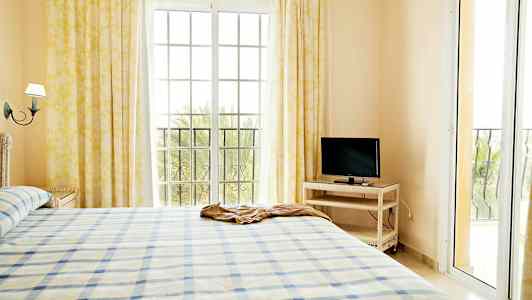 Valoisa makuuhuoneen sisustus, jossa on sänky, televisio pöydällä ja suuret ikkunat verhoilla. (Tekoälyn luoma teksti)