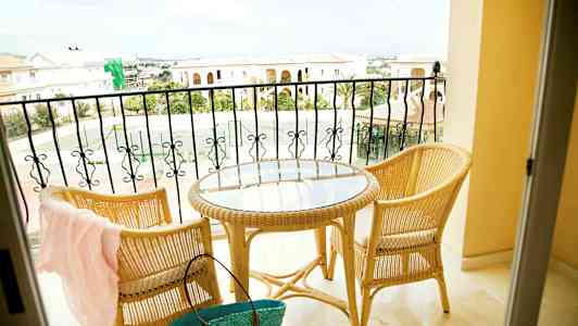 Kaksi rottinkituolia ja pyöreä lasipöytä parvekkeella, näkymä kivirakennuksiin ja tenniskenttään. (Tekoälyn luoma teksti)