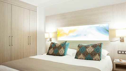 Hotellihuone, jossa on parivuode ja koristeelliset tyynyt, ympärillä vaaleaa puuta ja valaisimia. (Tekoälyn luoma teksti)