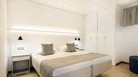 Kahden hengen huone, jossa valkoiset seinät, parisänky, sängyn lamput ja vaatekaappi. (Tekoälyn luoma teksti)