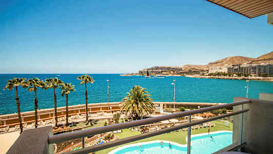 Näkymä parvekkeelta uima-allasalueelle ja rannikkolinjalle, jossa palmuja, meri ja rakennuksia kaukaisuudessa. (Tekoälyn luoma teksti)