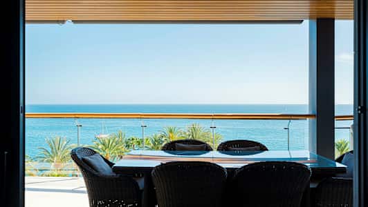 Terassi, jossa pöytä ja tuolit, merinäköala ja palmuja taustalla. (Tekoälyn luoma teksti)