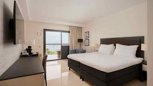 Hotellihuone, jossa suuri sänky, kirjoituspöytä ja televisio. Lasiovien takaa näkyy parveke ja merinäköala. (Tekoälyn luoma teksti)