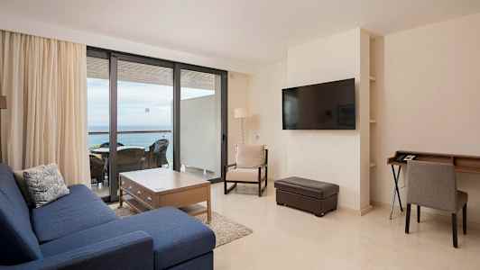 Olohuone, jossa sininen sohva, puinen pöytä ja tuoleja suuren ikkunan edessä parvekkeelle. (Tekoälyn luoma teksti)