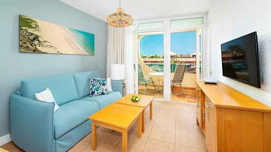 Olohuone, jossa on sininen sohva, kaksi puupöytää, televisio lipastolla ja näkymä terassille, jossa on ulkokalusteita. (Tekoälyn luoma teksti)