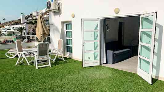 Ulkoalue, jossa vihreä nurmikko, istuinryhmä ja avoimet ovet sisätiloihin. (Tekoälyn luoma teksti)