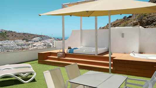 Terassi, jossa aurinkotuoleja, pöytä, varjo sekä näkymä mäkiseen ympäristöön rakennuksineen. (Tekoälyn luoma teksti)