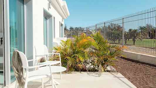 Terassi valkoisilla tuoleilla ja pöydällä, ympäröitynä vihreällä kasvillisuudella ja aidalla. (Tekoälyn luoma teksti)