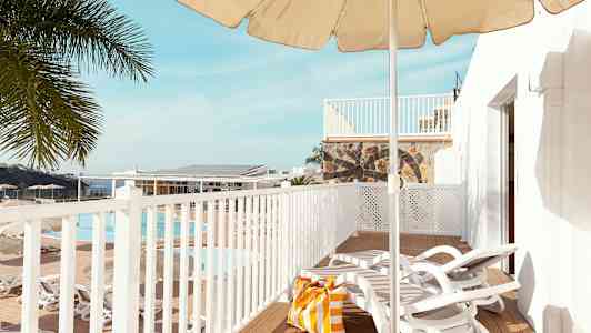 Näkymä terassille, jossa on aurinkotuoleja, aurinkovarjo ja rantakassi. Taustalla näkyy uima-allas ja palmuja. (Tekoälyn luoma teksti)