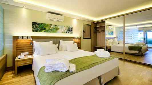 Hotellihuone, jossa on parisänky, vihreä huopa ja valkoiset tyynyt sekä taustalla peili ja vaatekaappi. (Tekoälyn luoma teksti)