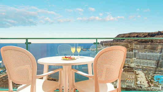 Kuva parvekkeesta, jolta on näkymä merelle ja ympäröiville rakennuksille. Parvekkeella on pöytä, jossa on kaksi lasia ja hedelmälautanen sekä kaksi tuolia. (Tekoälyn luoma teksti)